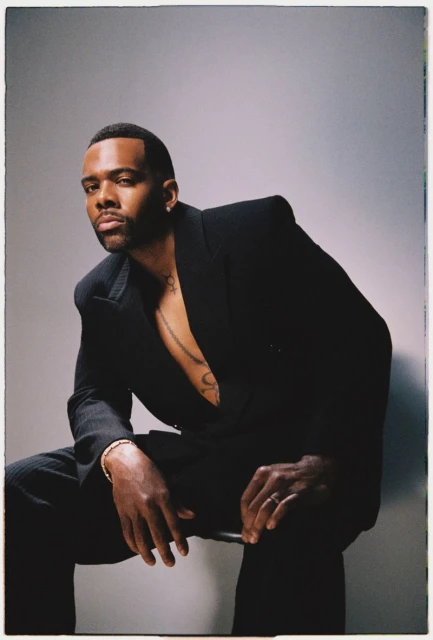 Portrait of an adult Black man with a beard sitting in a black suit and jewelry in a photo studio with gray background lighting.