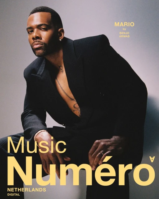 Portrait of African-American male singer Mario in a black suit, seated in a studio, for Numero magazine, bathed in soft studio lighting.