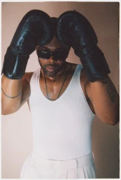 Black man wearing black boxing gloves, dark sunglasses, and a white ribbed tank top posing against a neutral backdrop.