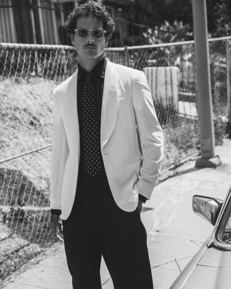 Black and white portrait of an adult male in a suit jacket and sunglasses, standing near a car on a city street.
