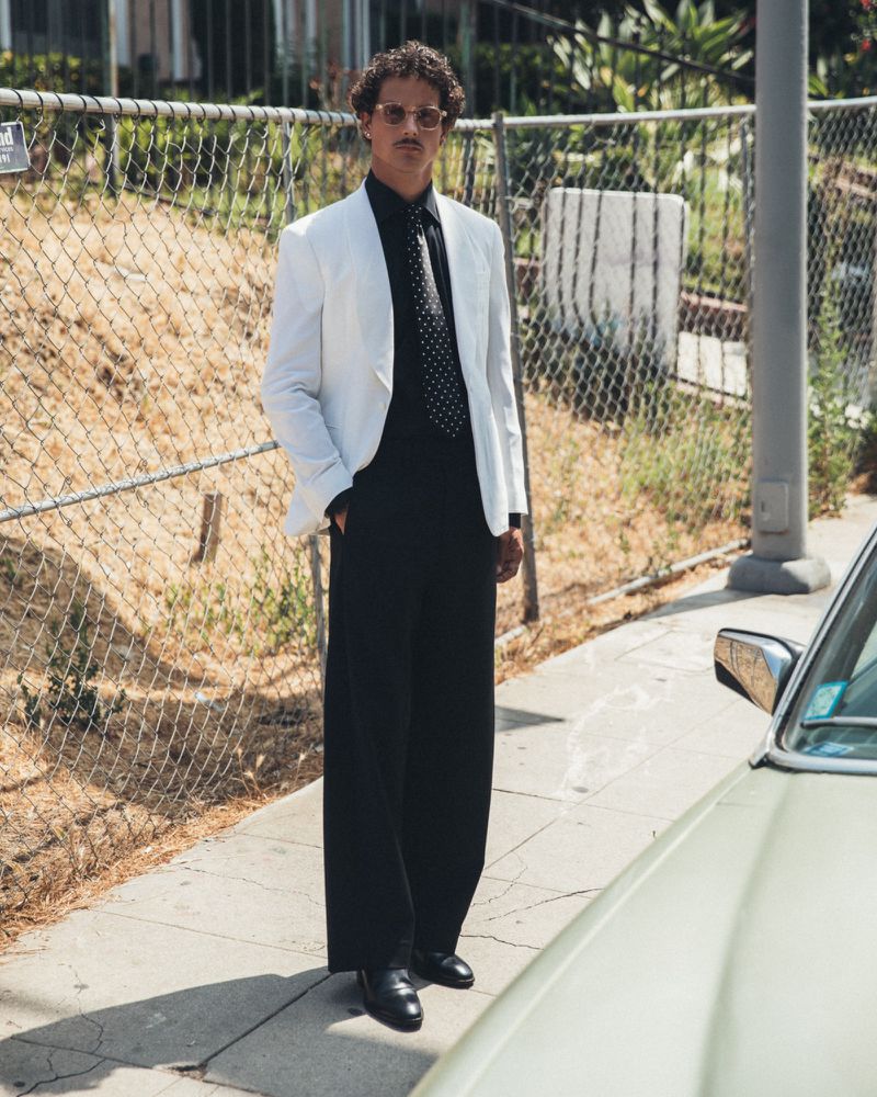 Stylish man in a white suit stands on a sidewalk next to a car and a metal fence in an urban environment, posing for a fashion shot.