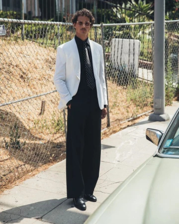 Stylish man in a white suit stands on a sidewalk next to a car and a metal fence in an urban environment, posing for a fashion shot.