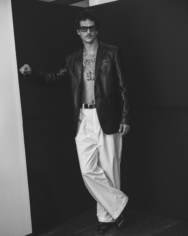 Studio shot of a man in a black leather jacket and white trousers, leaning against a black wall with a minimalist aesthetic.