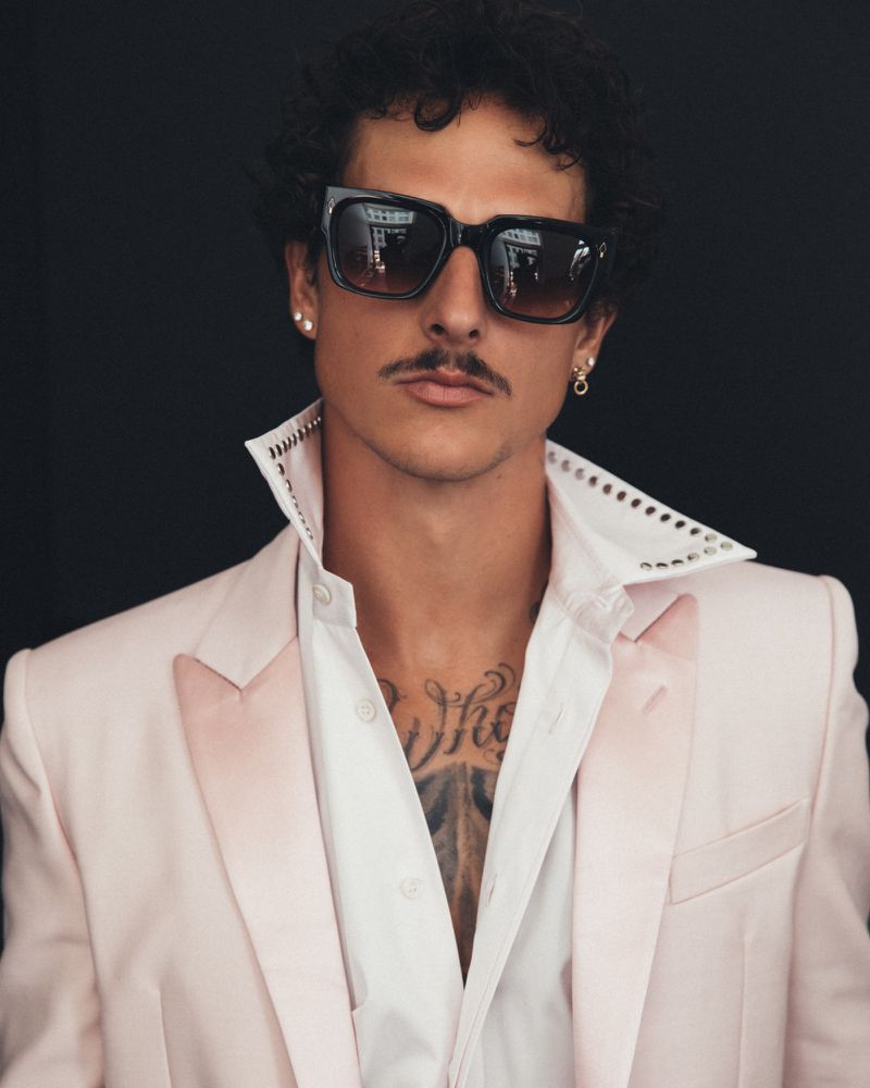Close-up studio portrait of a man with curly hair, a moustache, and sunglasses, wearing a pink blazer over a studded dress shirt.