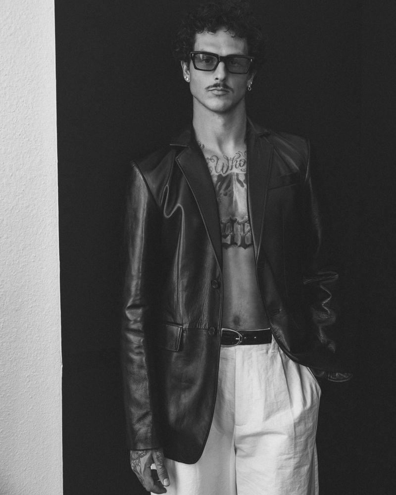Portrait of adult male with chest tattoos wearing a leather jacket and sunglasses against a dark background, captured in black and white.