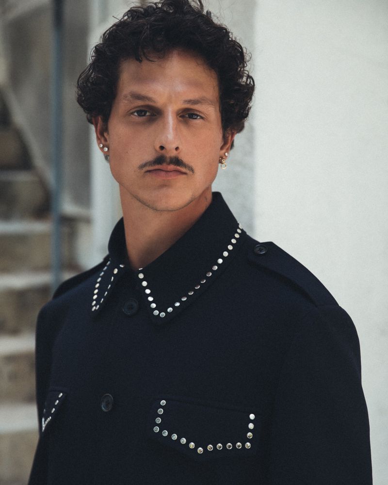 Portrait of a young adult male with curly hair and a mustache, wearing a dark blue jacket adorned with silver studs, posing indoors.