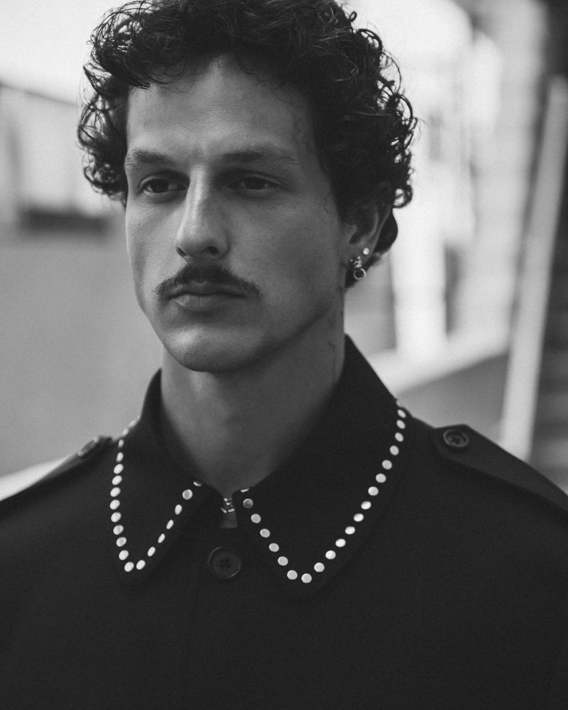 Monochrome portrait of an adult male with curly hair and a mustache, wearing a dark collared shirt with a studded design, neutral expression.