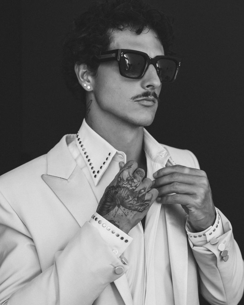 Black and white portrait of a man in sunglasses, wearing a white blazer, buttoning his shirt, against a dark background.