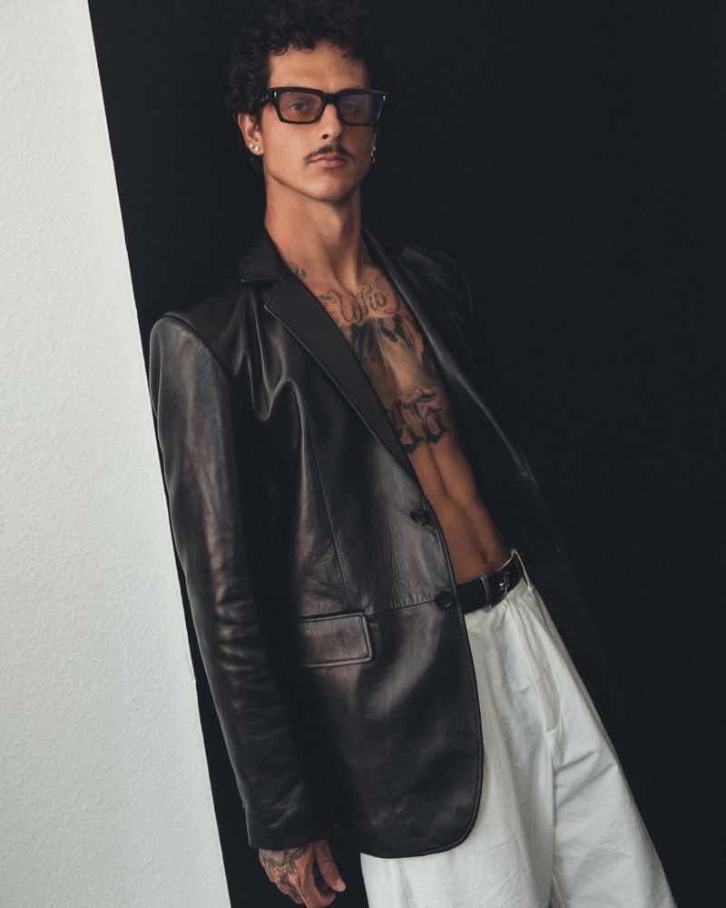 Portrait of a man wearing a leather blazer, glasses, and white pants against a black-and-white background in a studio setting.