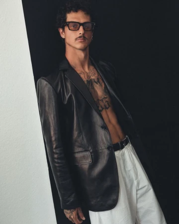 Portrait of a man wearing a leather blazer, glasses, and white pants against a black-and-white background in a studio setting.
