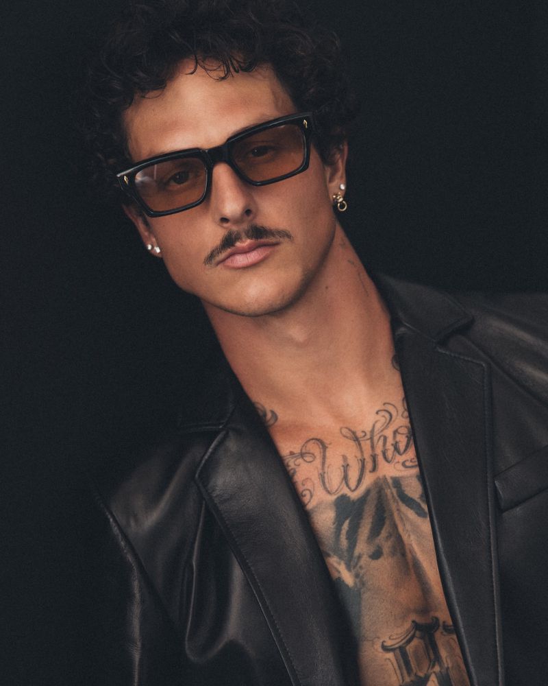 Nick Mayhugh wearing tinted rectangular sunglasses and an open black leather jacket showing his tattooed chest against a black background.