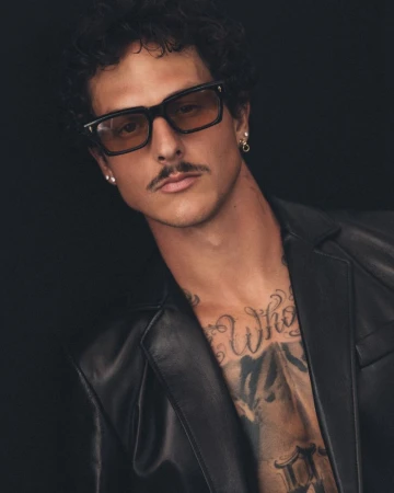 Nick Mayhugh wearing tinted rectangular sunglasses and an open black leather jacket showing his tattooed chest against a black background.