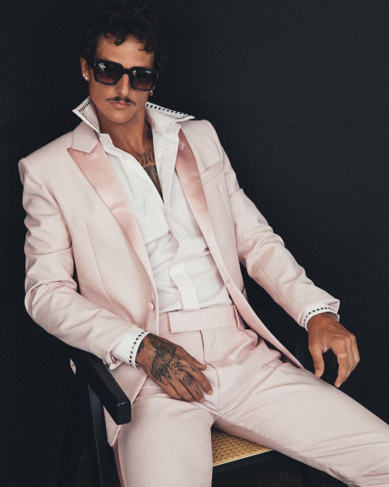 Stylish man in a light pink suit poses in a studio with a dark background, showcasing fashion and sophistication.