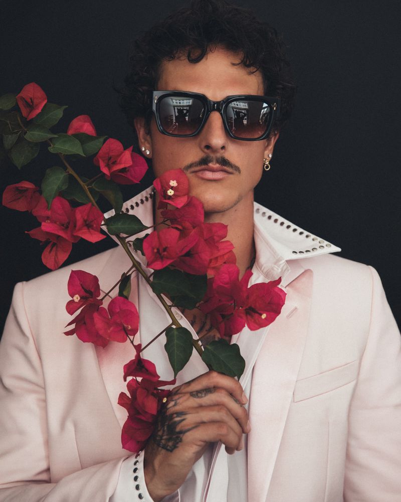 Stylish adult male in sunglasses and a pink blazer, holding a branch with red bougainvillea flowers against a dark background.