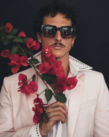 Stylish adult male in sunglasses and a pink blazer, holding a branch with red bougainvillea flowers against a dark background.