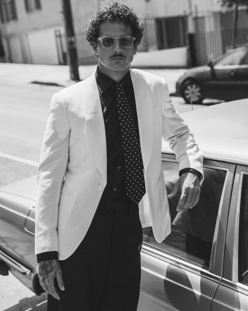A man in a suit and tie stands in front of a vintage car in black and white, posing for a fashion editorial, and styled with retro sunglasses.