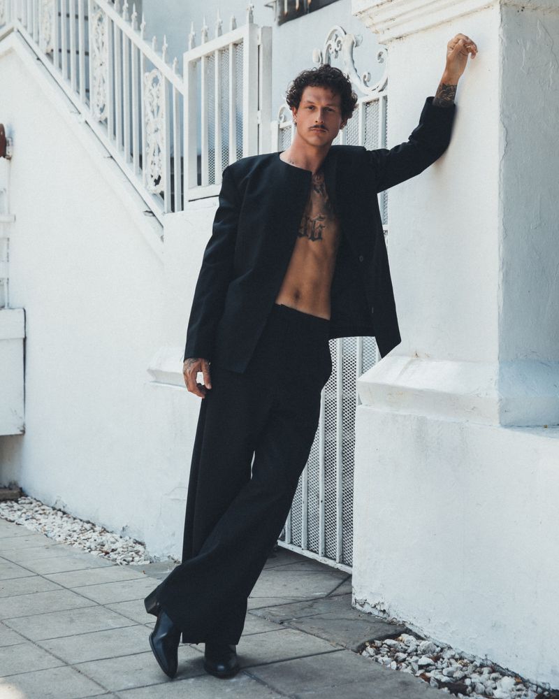 Nick Mayhugh wearing an open black suit jacket with no shirt and wide-leg trousers while leaning against a white exterior wall.