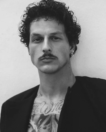 Black and white portrait of Nick Mayhugh with curly hair, a mustache, and chest tattoos wearing an open black jacket.