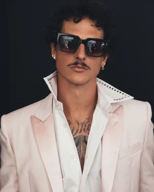 Nick Mayhugh wearing thick black sunglasses, an open-collar white shirt, and a pale blush blazer against a black studio backdrop.