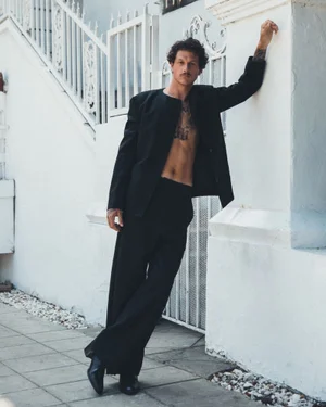 Nick Mayhugh wearing an open black suit jacket with no shirt and wide-leg trousers while leaning against a white exterior wall.