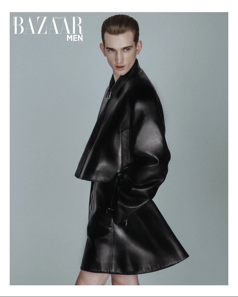 Fashion editorial shot of a red-haired male model in a black leather jacket and skirt against a grey background for Bazaar Men magazine.