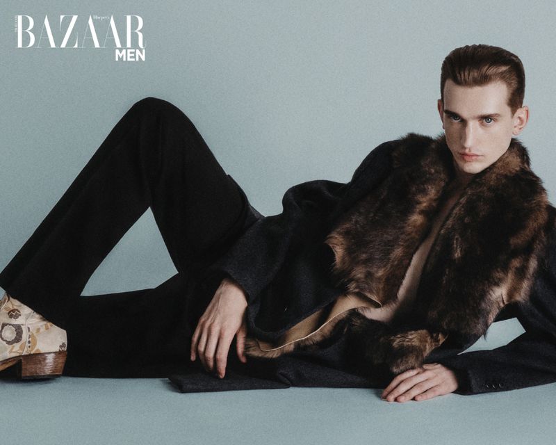 Male fashion model laying down wearing an overcoat with fur collar and patterned dress shoes in a studio fashion shot.