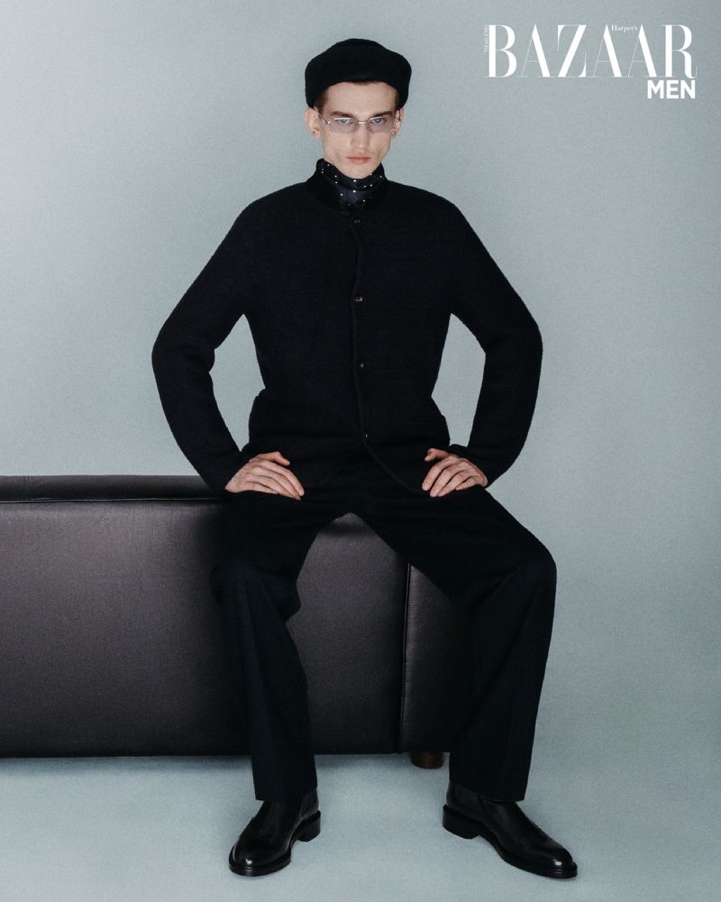 Elegant man in all-black attire and glasses seated on a dark couch against a plain backdrop in a studio setting. Fashion editorial.