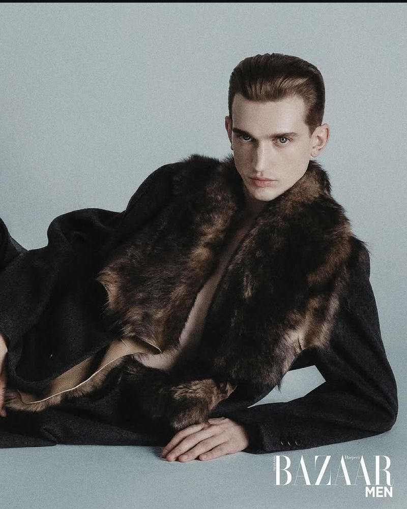 Fashion shot of male model in dark coat with large fur collar lying down looking at the camera in a light gray studio.