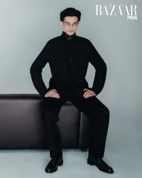 Model Eugen Ivanov seated on a dark leather bench wearing an all-black suit, beret, and glasses for Harper's Bazaar Men.