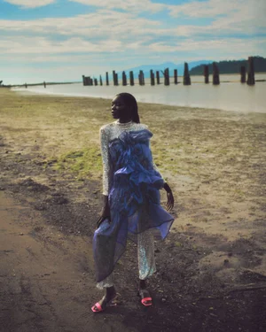 Model wearing sequined outfit with blue ruffled overlay standing on a sandy beach by water with wooden pilings