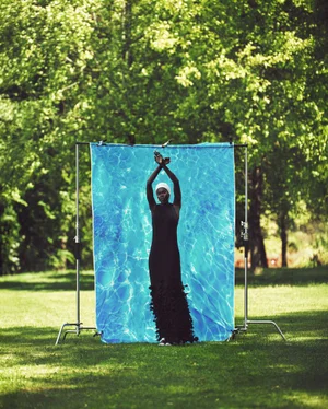 Model in a black maxi dress and white cap posing before a water print backdrop outdoors on a green lawn.