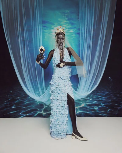 Black model in a light blue petal-embroidered halter gown with beaded fringe headdress standing before an underwater backdrop.