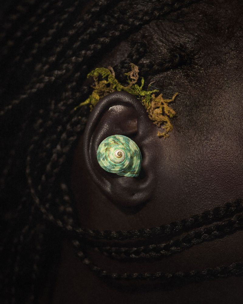 Close-up of an adult female with dark skin, wearing a shell earring and some greenery behind her ear, against a dark background.