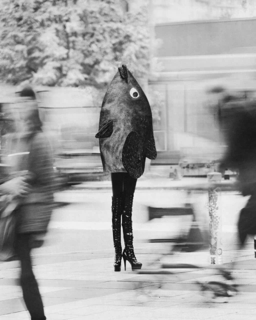 Person in fish costume and platform boots stands on city street with blurred passersby, monochrome image.