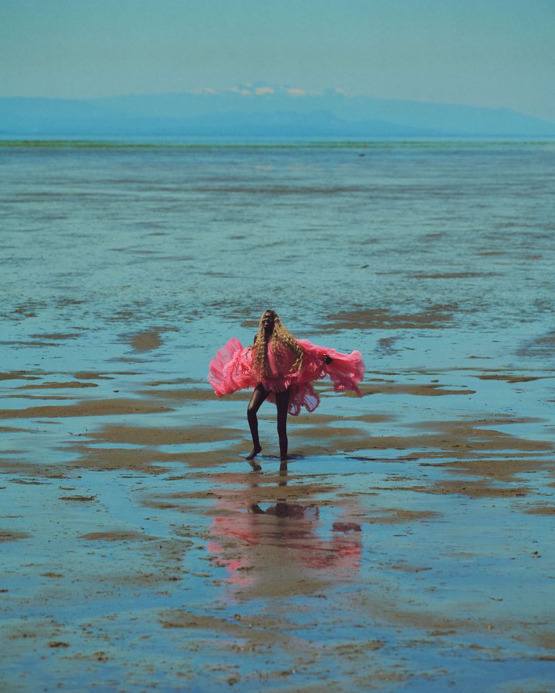 A woman in a pink dress dances barefoot on a wet beach with mountains in the distance, creating a sense of freedom and artistry.