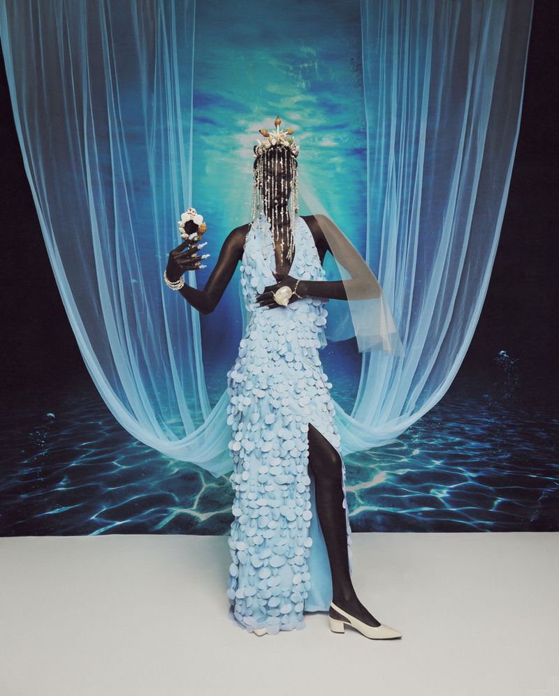 A model in an embellished dress poses with a seashell in front of an underwater-themed backdrop, wearing a beaded headdress and white heels.