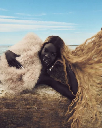 A striking photograph features a woman with long, wavy hair and dark skin, lying on a wooden log by the sea under a blue sky.