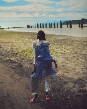 A model poses in a glittering outfit and pink heels on a sandy beach with wooden pilings in the water and mountains in the distance.