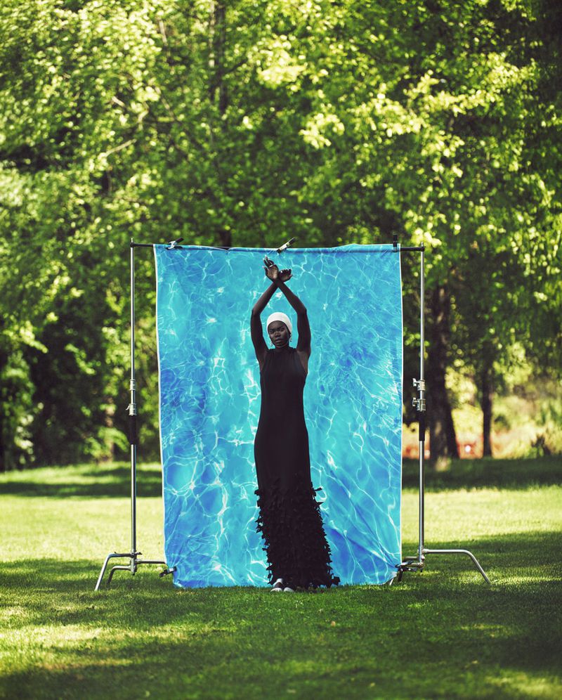 Woman in black dress poses in front of blue water-patterned backdrop on green lawn, surrounded by trees and photography equipment.