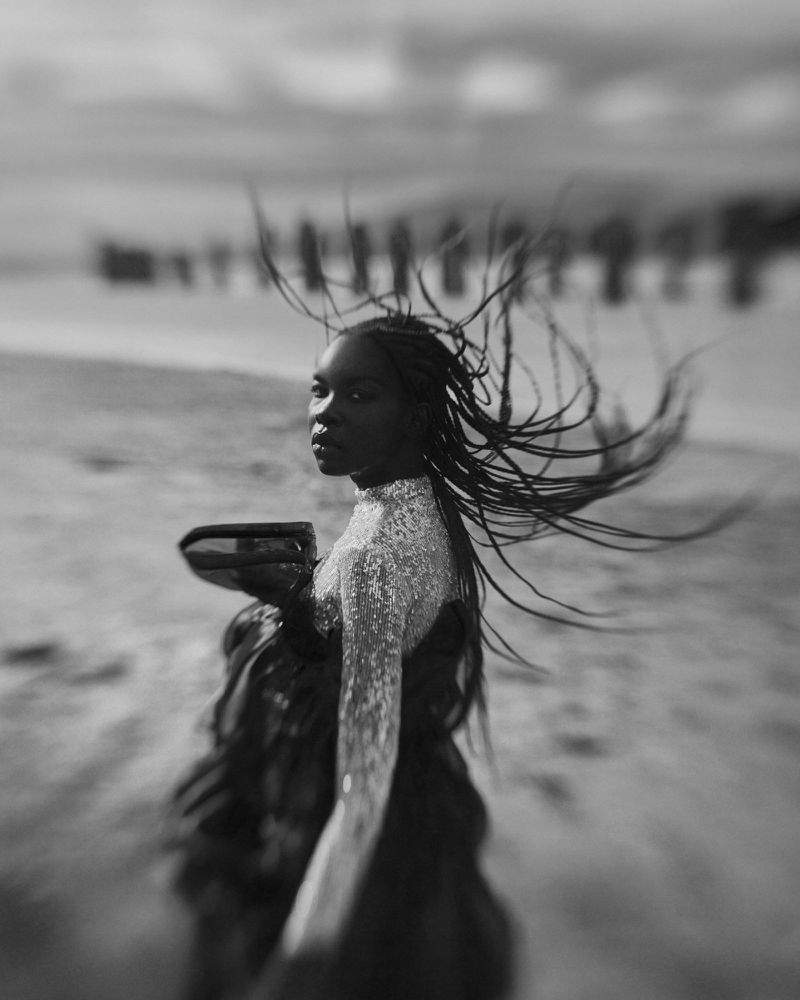 Monochrome portrait of an African woman with flowing braided hair, wearing a dress at the beach, with a shallow depth of field.