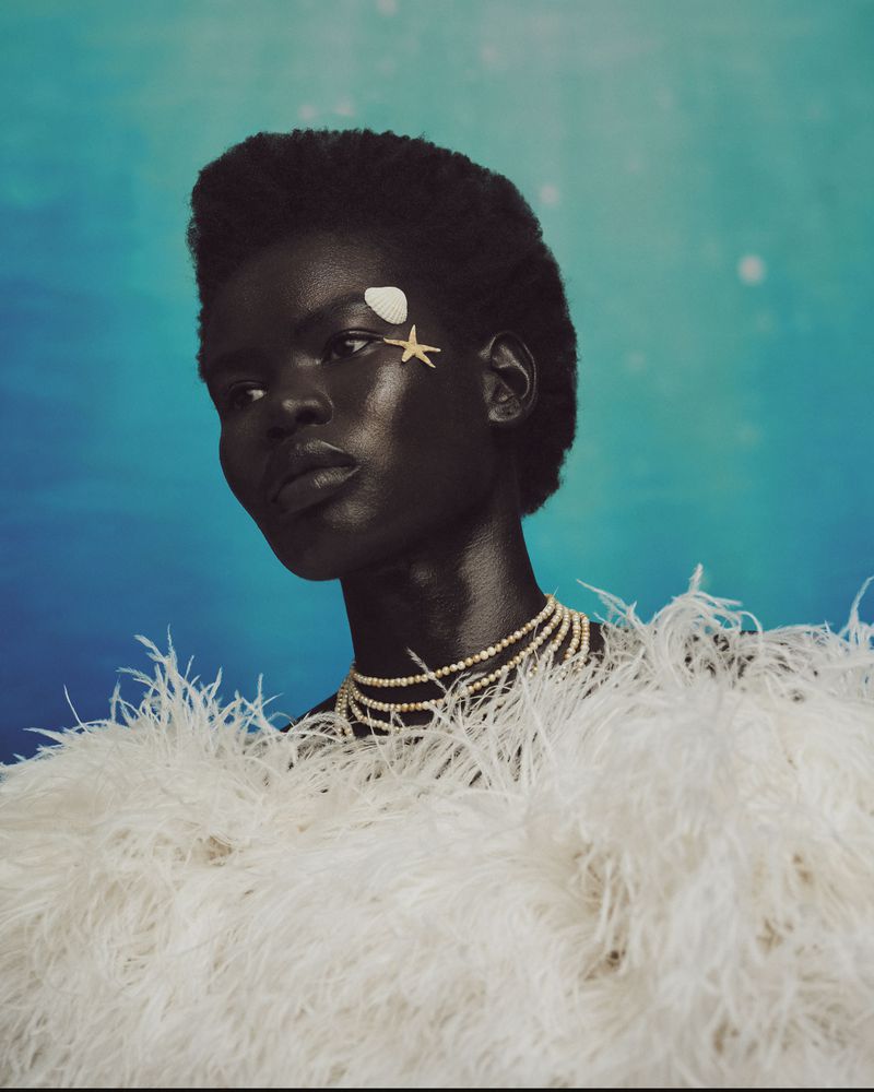 Portrait of an African woman with seashell and star accessories, white feather top, set against a blue watercolor background.