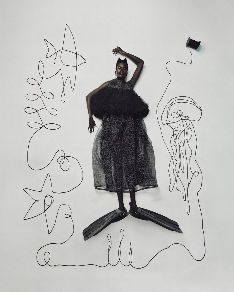 Fashionable Black model poses with fins in an abstract art studio. She wears a black dress and stands against a white backdrop.