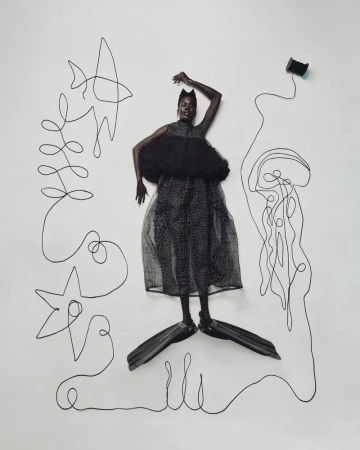 Fashionable Black model poses with fins in an abstract art studio. She wears a black dress and stands against a white backdrop.
