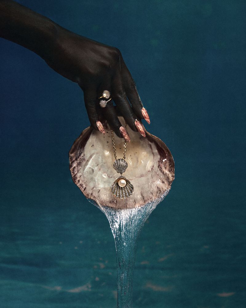 Hand with dark painted skin holds a shell with a pearl necklace, and a pearl ring, with water flowing from the shell in studio shot.