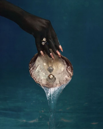 Hand with dark painted skin holds a shell with a pearl necklace, and a pearl ring, with water flowing from the shell in studio shot.