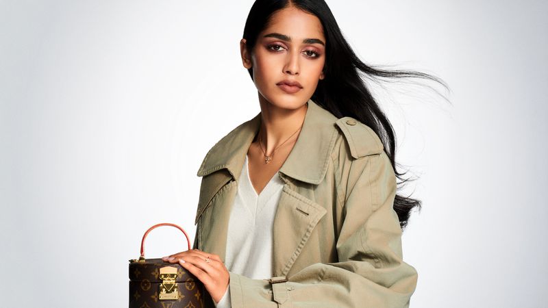 Adult woman with long dark hair poses with a brown Louis Vuitton trunk bag in front of a white background in a studio setting.