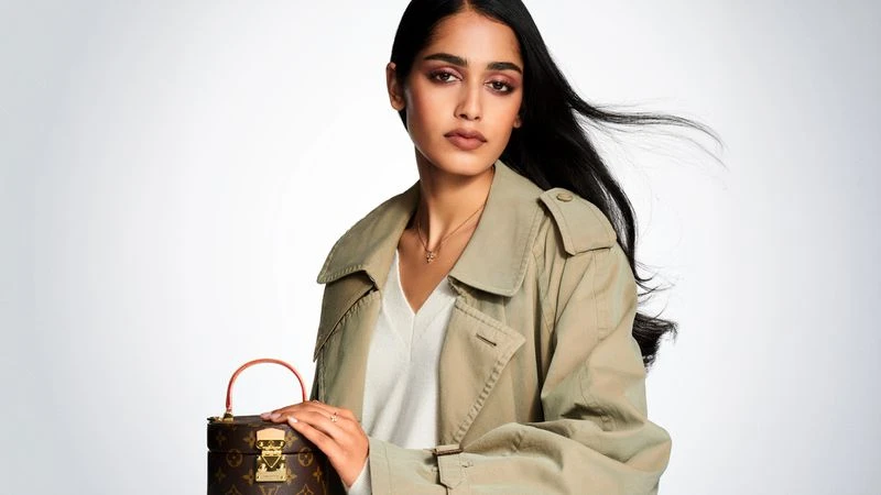 Adult woman with long dark hair poses with a brown Louis Vuitton trunk bag in front of a white background in a studio setting.
