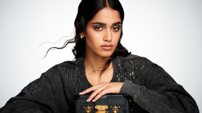 A young woman with dark hair poses indoors in a black denim jacket, holding a black Louis Vuitton trunk against a bright white background.