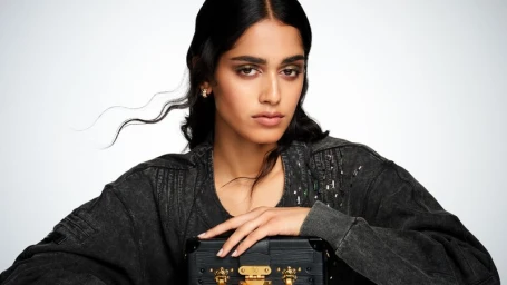 Model Mahi Kabra in a washed charcoal jacket holding a black Louis Vuitton trunk clutch against a clean studio backdrop.