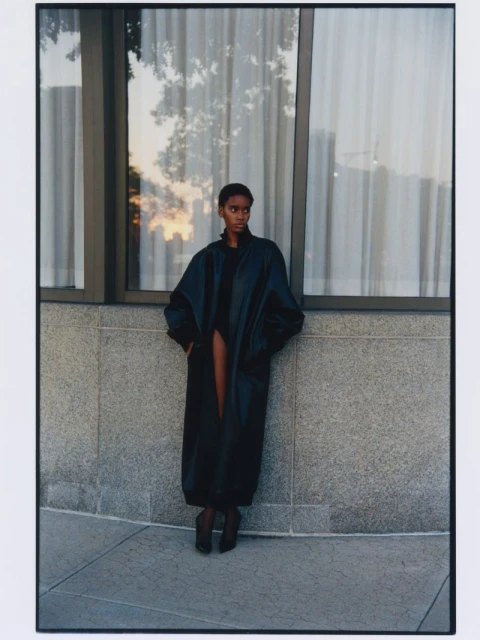 A woman in a black coat stands outdoors against a building facade with large windows reflecting the setting sun, creating an urban fashion scene.